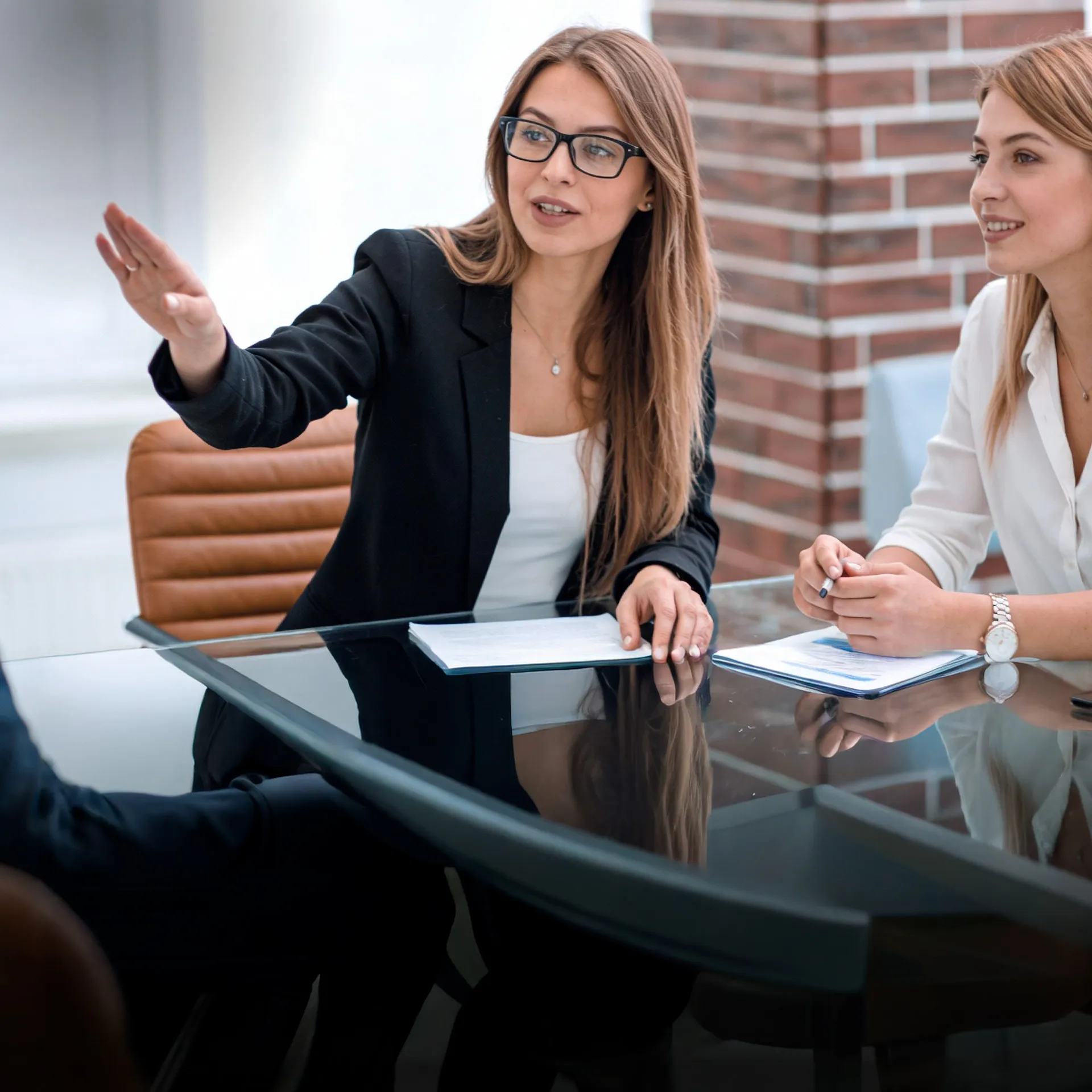 Two businesswomen sit at a glass conference table; the woman in a black blazer gestures as she talks to her colleague next to her.