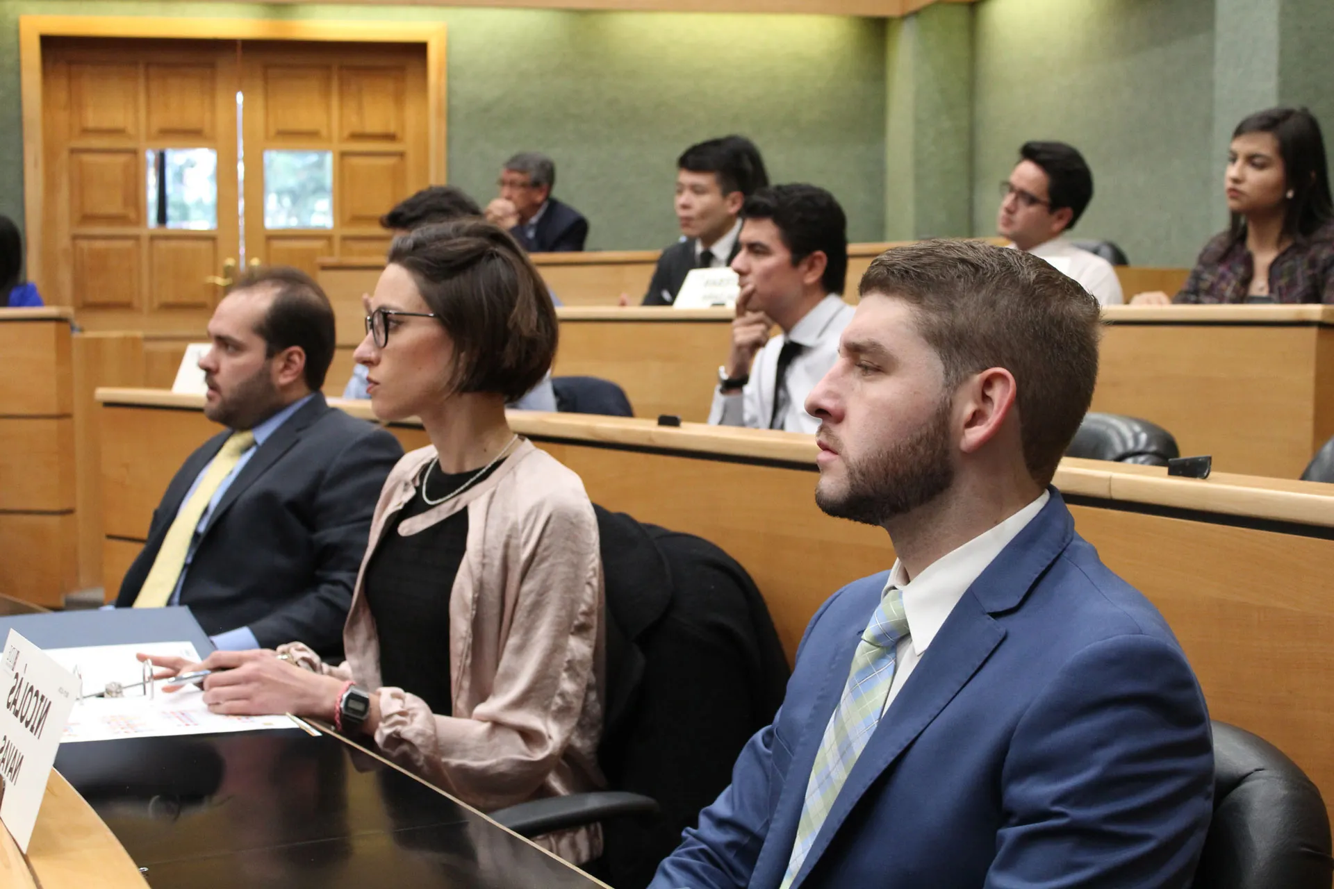 Audience members in a formal meeting room: man in blue suit and striped tie, woman with glasses and beige jacket in the foreground.