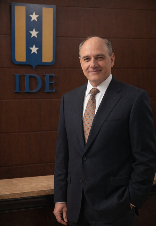 Businessman in a dark suit and patterned tie stands in front of a wood-paneled wall with an IDE logo behind him.