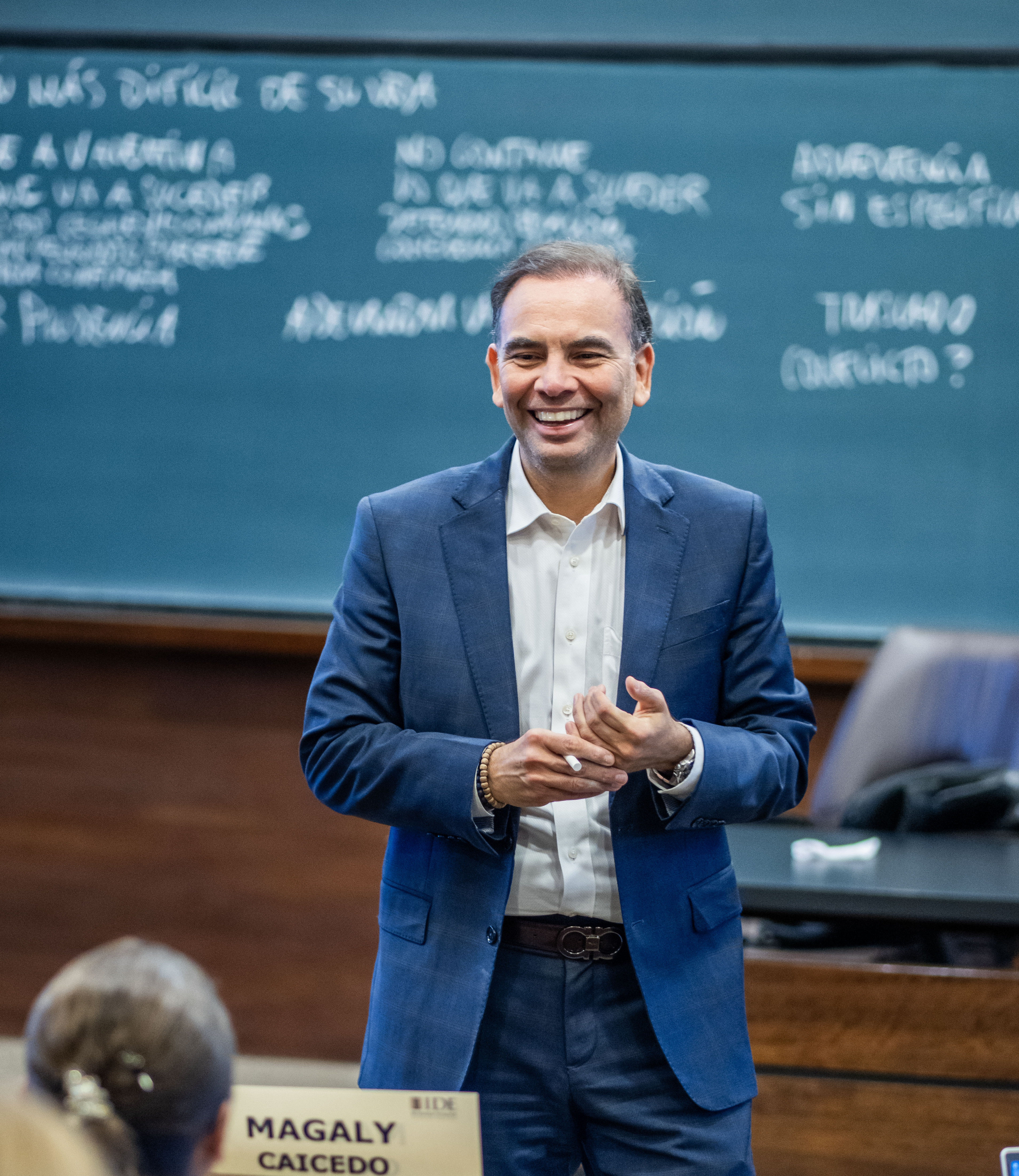 Smiling man in a blue suit giving a talk in a lecture hall, chalkboard behind him.